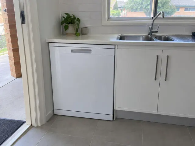 White dishwasher installed under a kitchen benchtop beside a sink and cabinets in a tiled home kitchen in Telarah